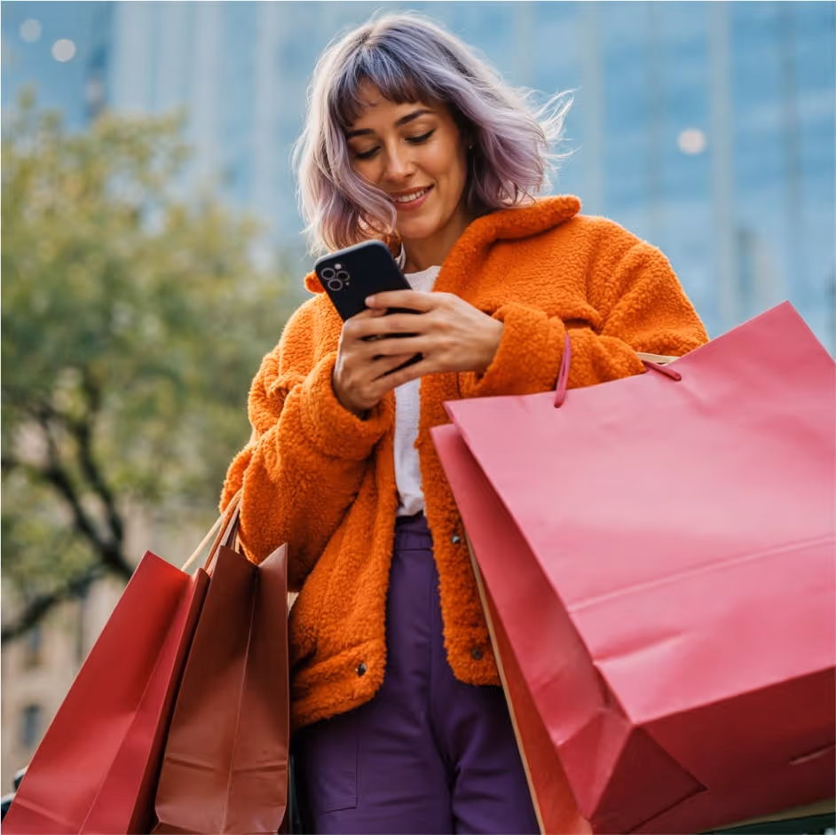 Pessoa de casaco laranja olhando o celular com sacolas de compras na mão